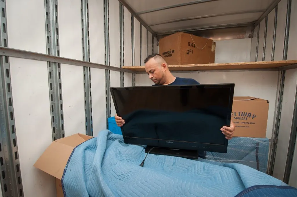 Two Beltway movers lifting a large flat-screen television over a padded surface in a living room